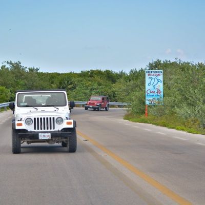 Two Jeeps on a road near greenery and a welcome sign on a clear day.