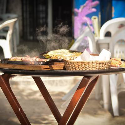 Outdoor table with a steaming food platter and basket on a wooden stand near white chairs.