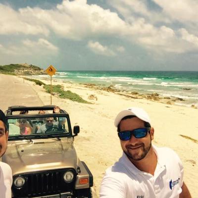 Two men take a selfie with an open-top jeep by the beach under a partly cloudy sky.