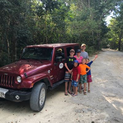 Family of six posing by a red Jeep on a forest dirt road.