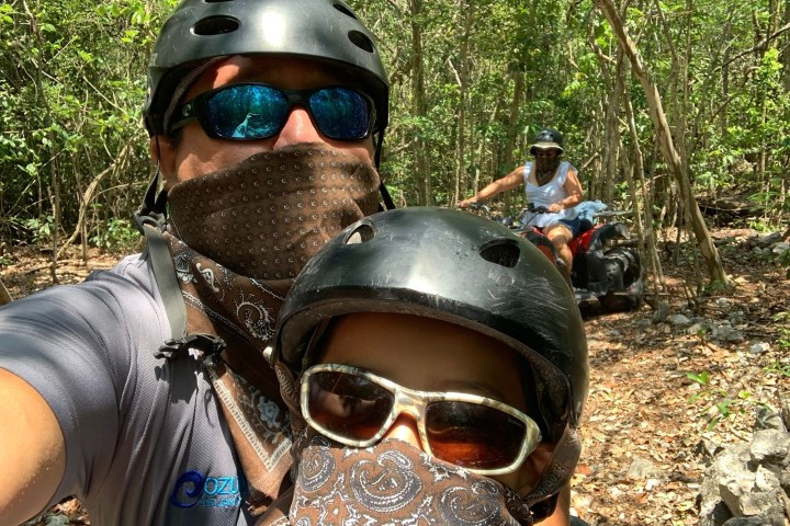 People with helmets and bandanas on an ATV ride through a wooded area.