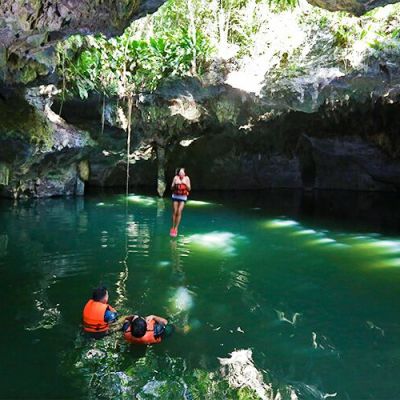People swimming in a sunlit cave pool with lush greenery above.