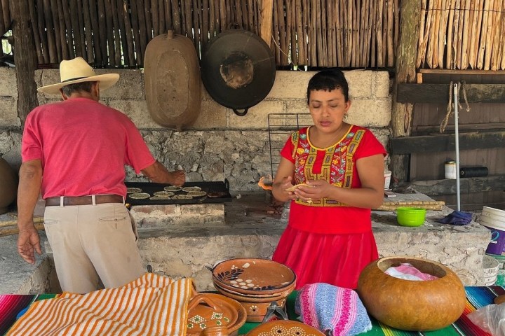 Two people preparing traditional food in an outdoor kitchen with clay pots and colorful fabrics.