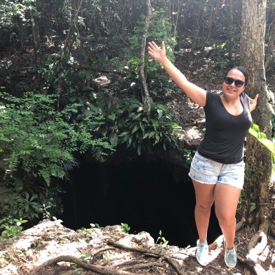 Smiling woman posing beside a tree in a lush forest with her arms raised.