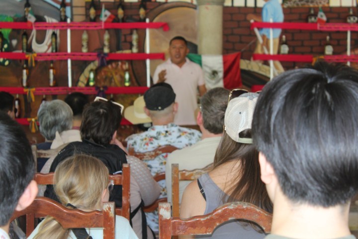 Audience listening to a speaker indoors with Mexican flag in the background.