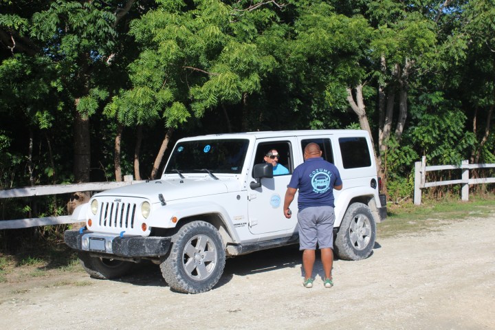 White Jeep stopped on a dirt road, person standing and talking to driver, trees in background.