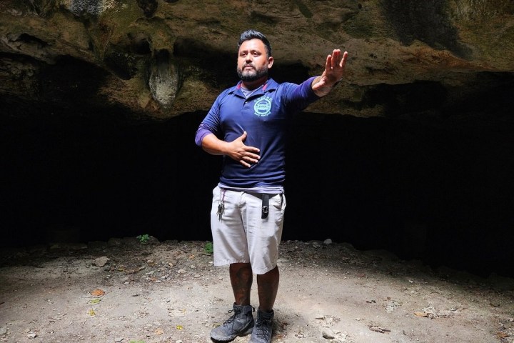 A man stands in a cave, gesturing with one hand, wearing shorts and boots.